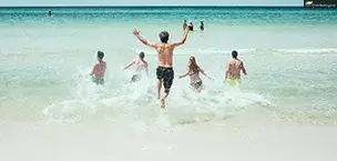 Cruise_Holidays A group of people running into the ocean on a sunny beach day, with clear blue water and sky.