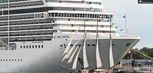 Cruise_Holidays A large cruise ship, MSC Poesia, docked with a smaller sailboat in front at a port.