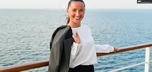 A woman smiles on a cruise ship deck, holding a business jacket, with the ocean in the background.