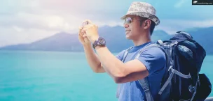 First_Time_Cruisers Traveler in a hat and sunglasses taking photos by the sea with scenic mountains in the background.