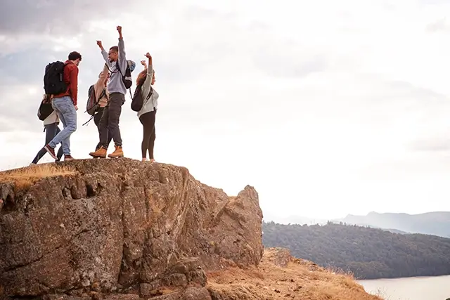 At the summit of a mountain, five young adult friends cheer with their arms up in the air
