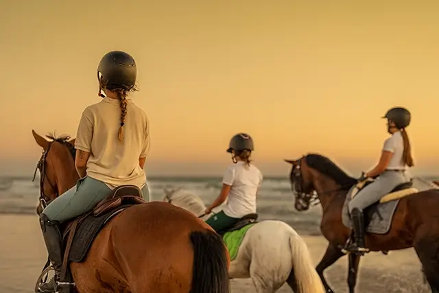 A young woman with braids is riding a horse along with two other young riders on the beach