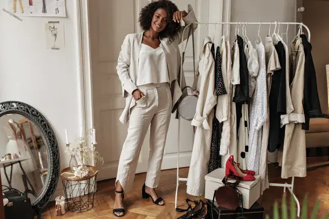 This full-length portrait shows a stylish dark-skinned curly woman in white pants and top leaning on a hanger in the dressing room