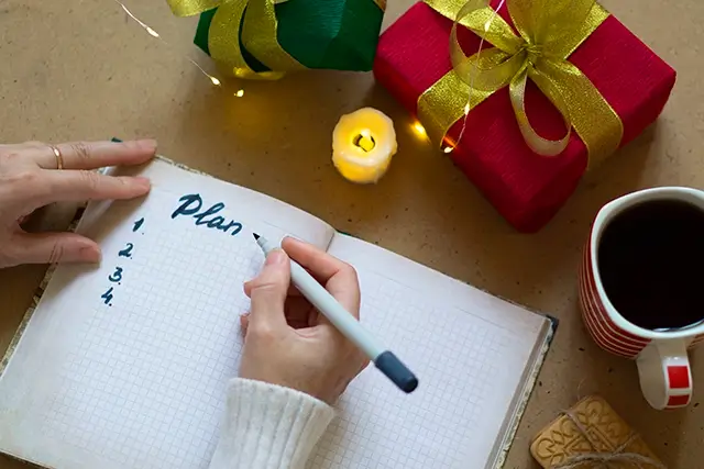 Writing in the planner is a woman's hand in a white sweater. Photo with selective focus, bokeh lights, and atmospheric effects. Gifts and shopping concept for Christmas
